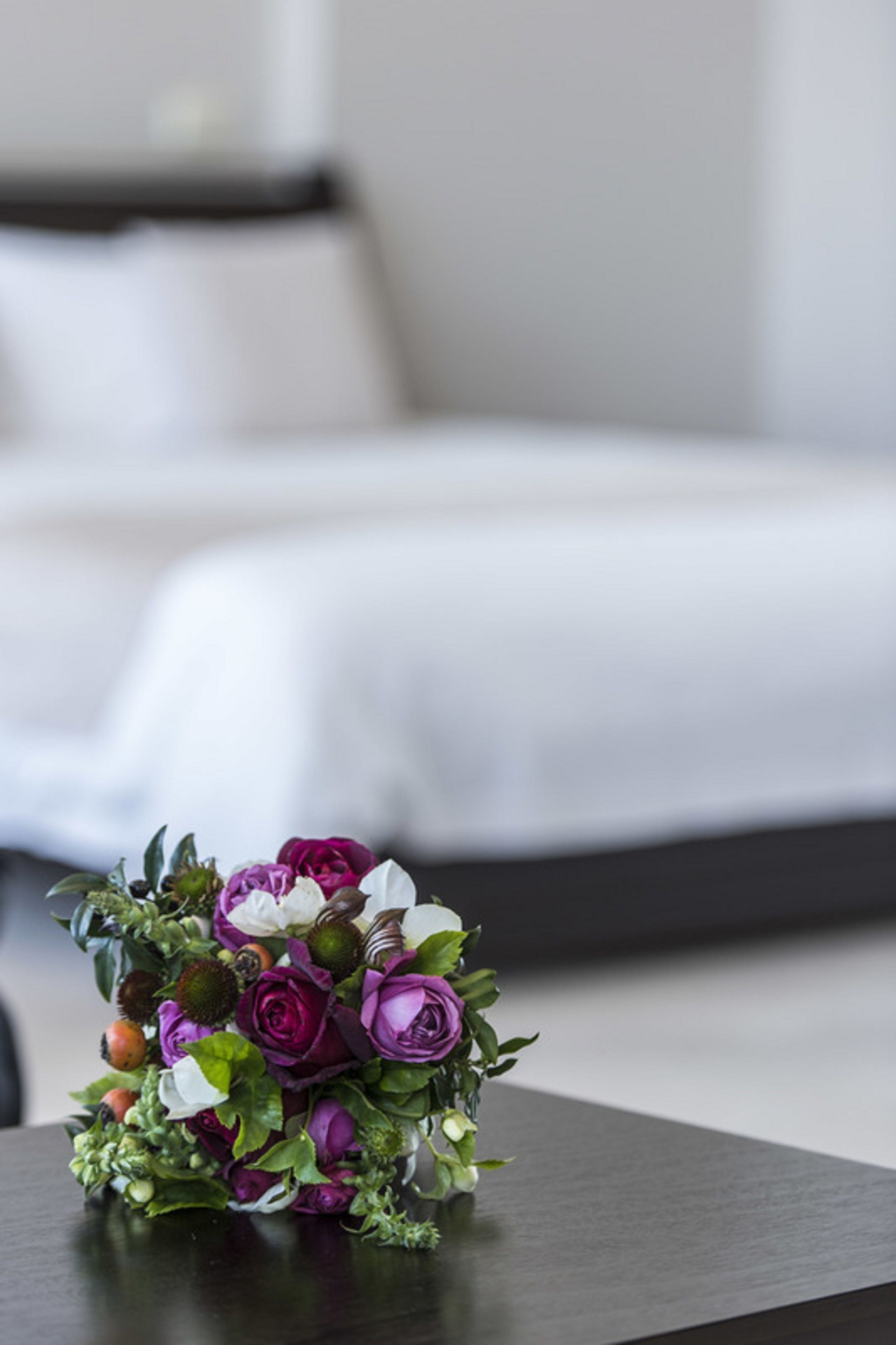 A bouquet of purple and white flowers on a dark table in a room with a white bed and a dark nightstand.