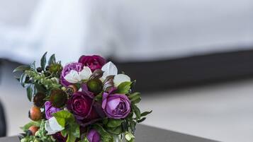 Un ramo de flores moradas y blancas sobre una mesa oscura en una habitación con una cama blanca y una mesita de noche oscura.