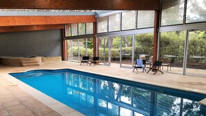 Una piscina interior con paredes de vidrio transparente, techo de madera y zona de patio con sillas y un jacuzzi.