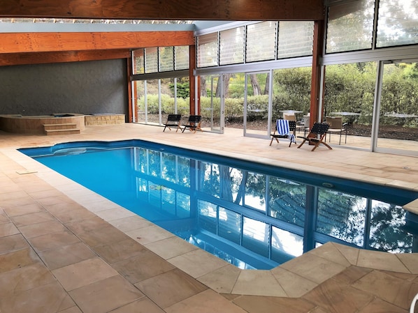 An indoor swimming pool with clear glass walls, a wooden ceiling, and a patio area with chairs and a hot tub. - Macedon Ranges Hotel & Spa (Macedon)