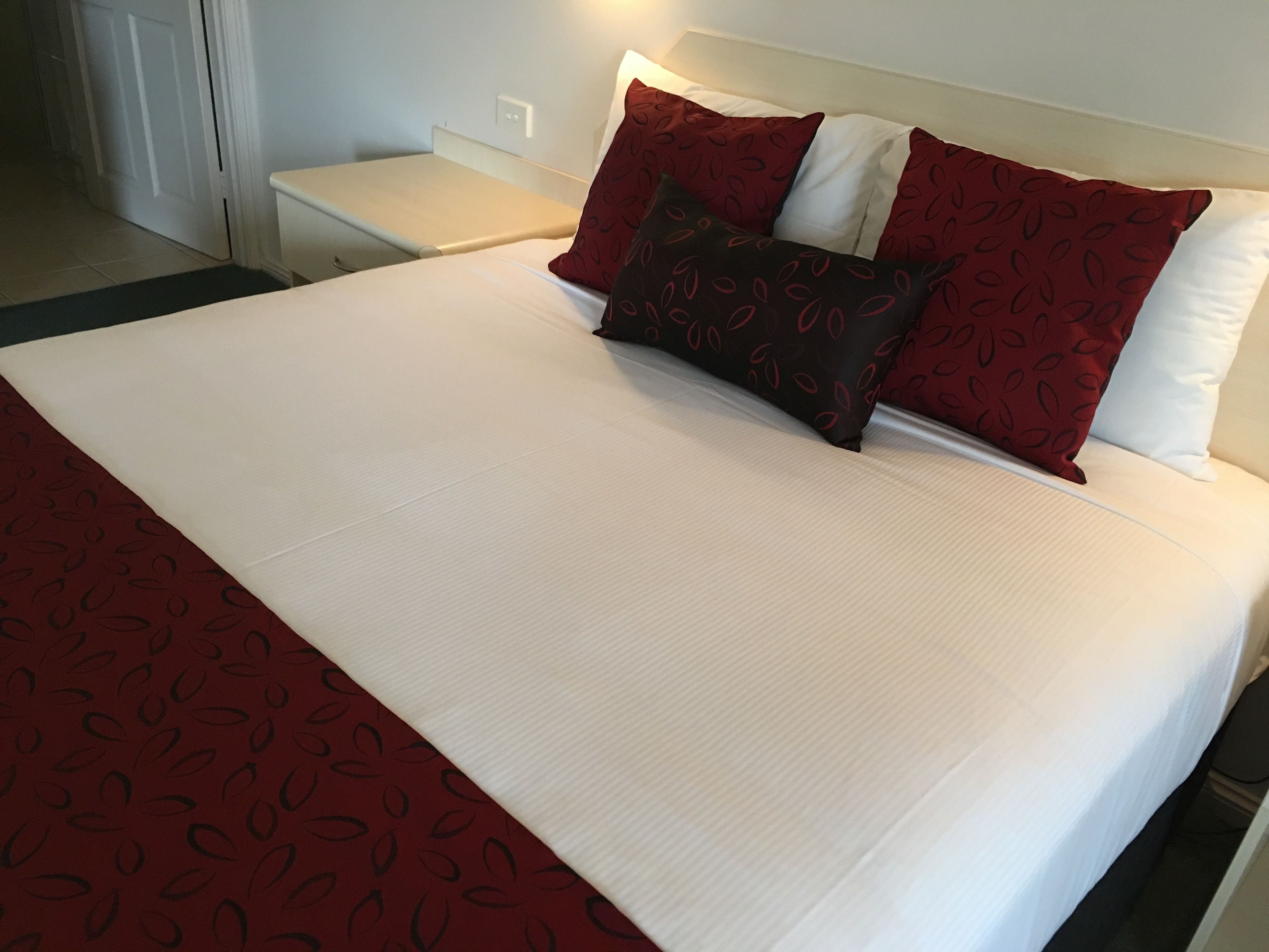 Ein ordentlich bezogenes Bett mit weißer und roter Bettwäsche, ein dunkelbraunes Kopfkissen mit Blätermuster und eine rote und weiße Tagesdecke.