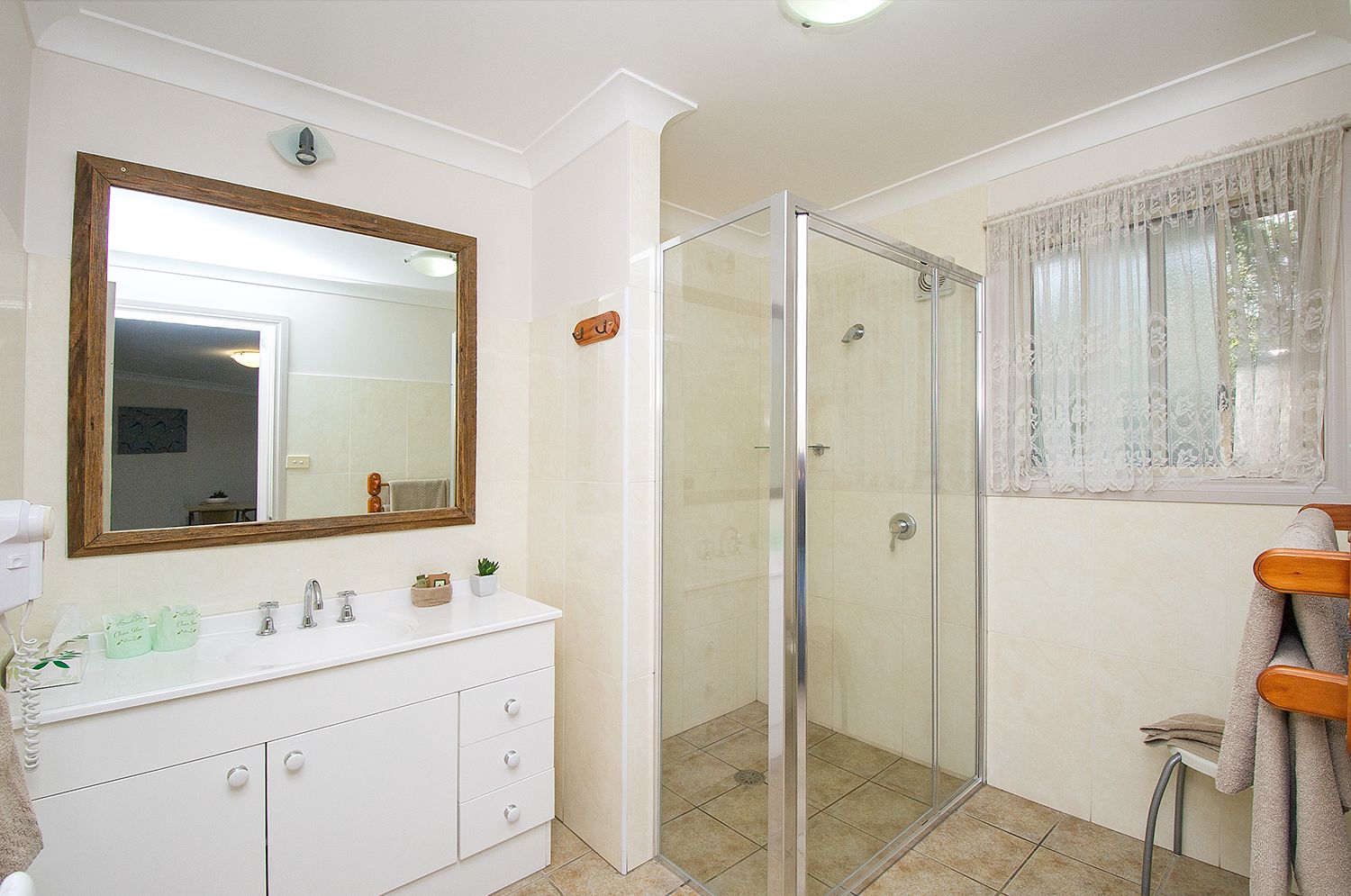 A bathroom with a white vanity, a large mirror, a glass shower enclosure, and a window with lace curtains.