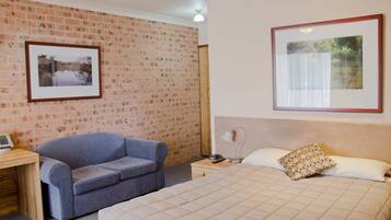 Un dormitorio con pared de ladrillo, cama con cubrecama acolchado, sofá azul, mesita de noche con lámpara y cuadros en la pared.