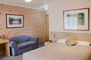 A bedroom with a brick wall, a bed with a quilted cover, a blue sofa, a bedside table with a lamp, and framed pictures on the wall.