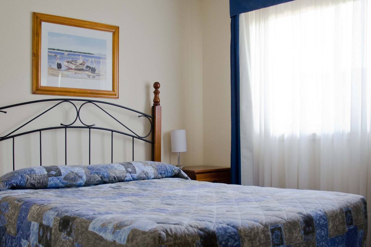 A neatly made bed with a quilt, a ceiling fan, a framed picture on the wall, and a window with curtains.