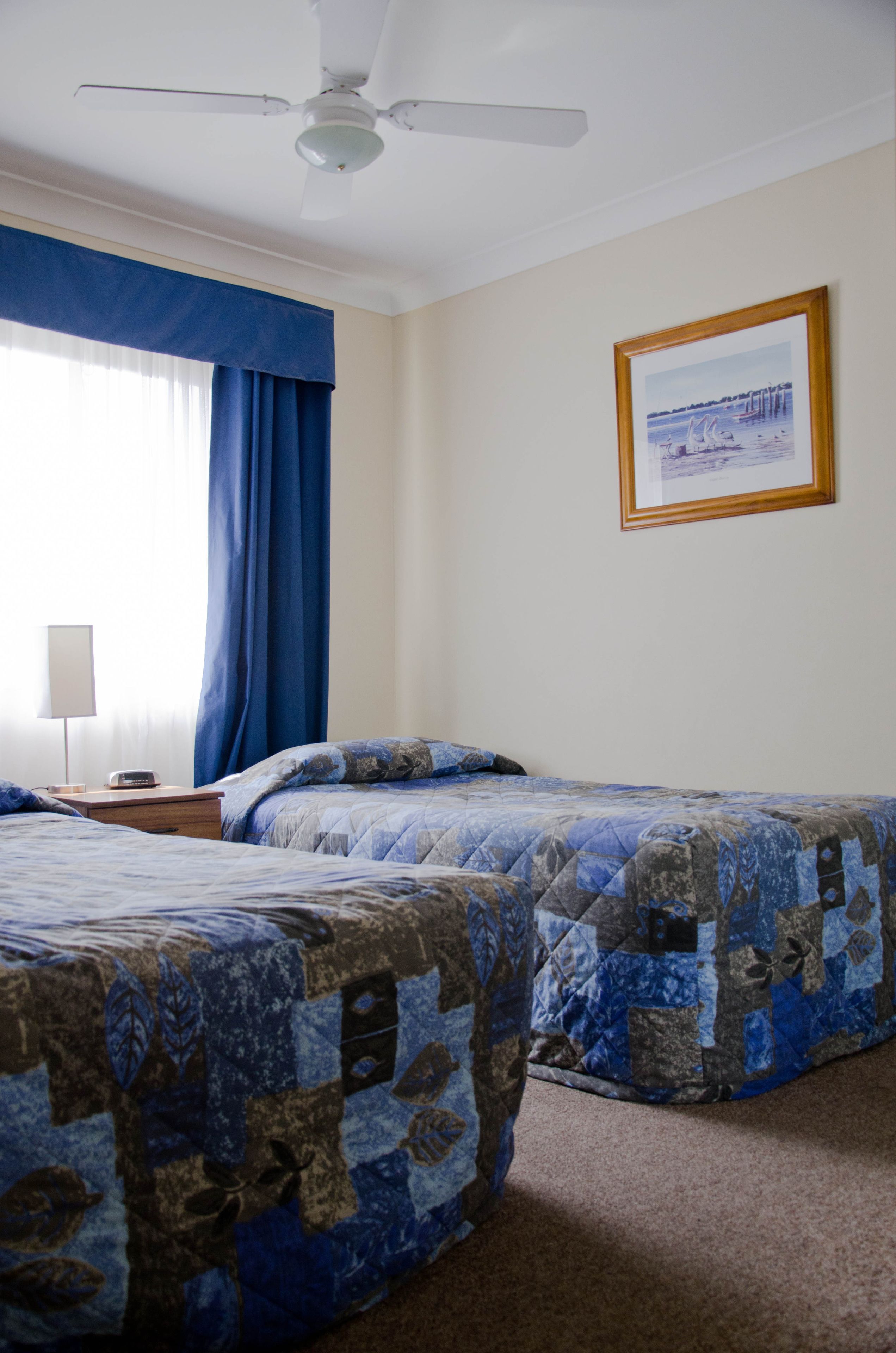 Habitación de hotel con dos camas, un ventilador de techo, un cuadro enmarcado en la pared y una ventana con cortinas azules.