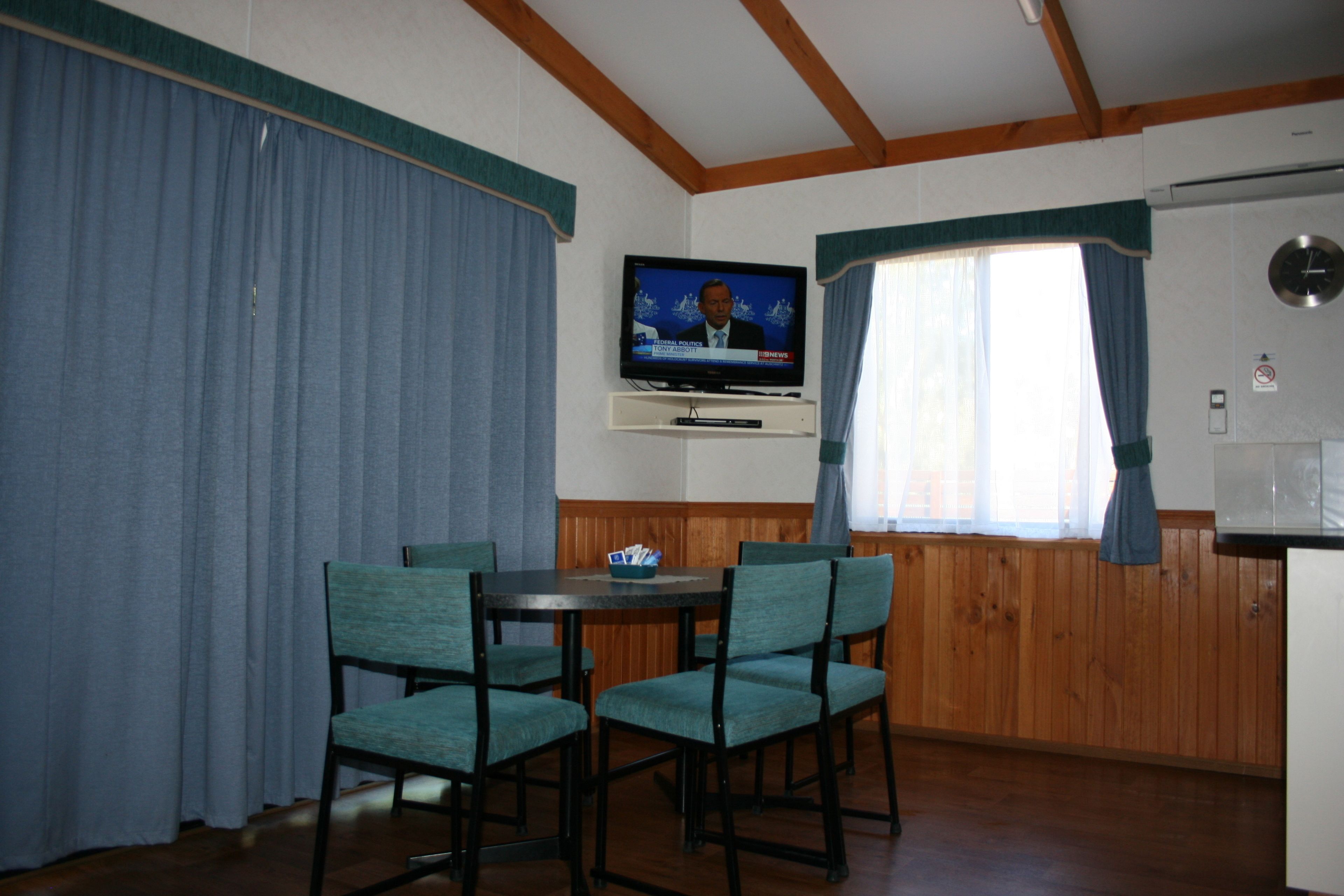 A room with a television mounted on the wall, a dining table with chairs, wooden paneling, and a window with curtains.