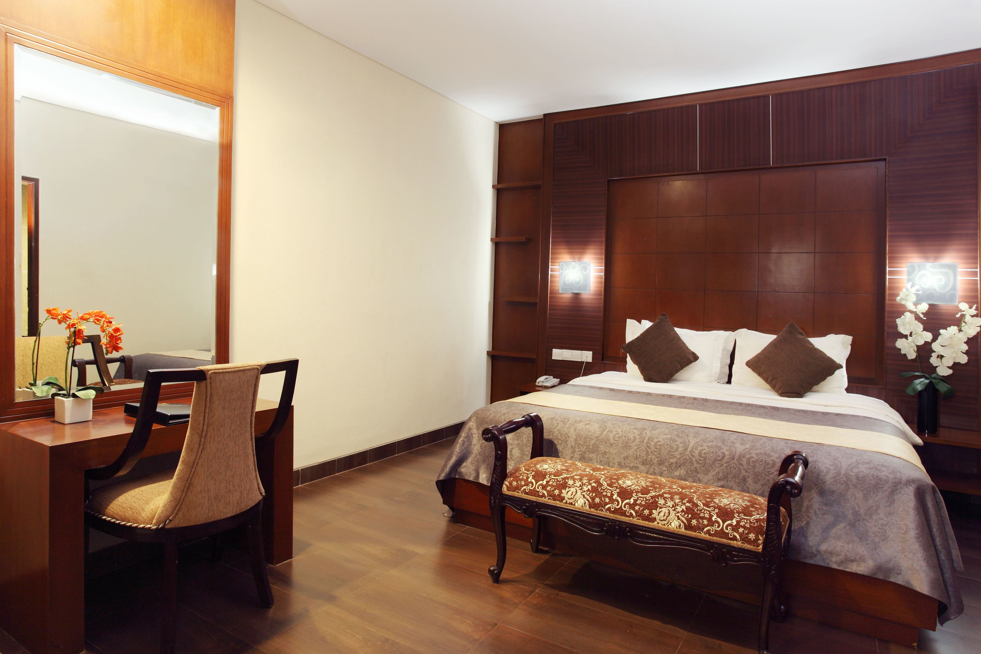 A hotel room with a wooden bed, a desk with a chair, a bench, and a large mirror.