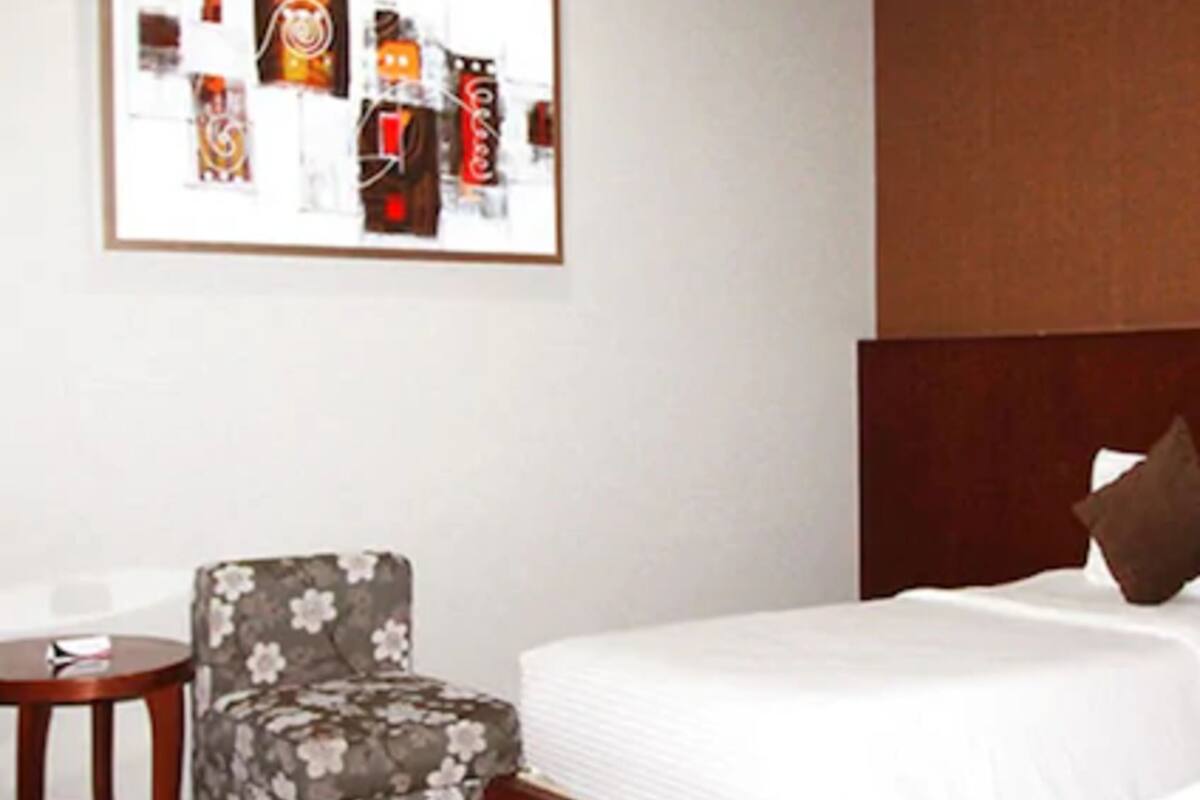 A hotel room with a bed, a chair, a small table, and a framed picture on the wall.