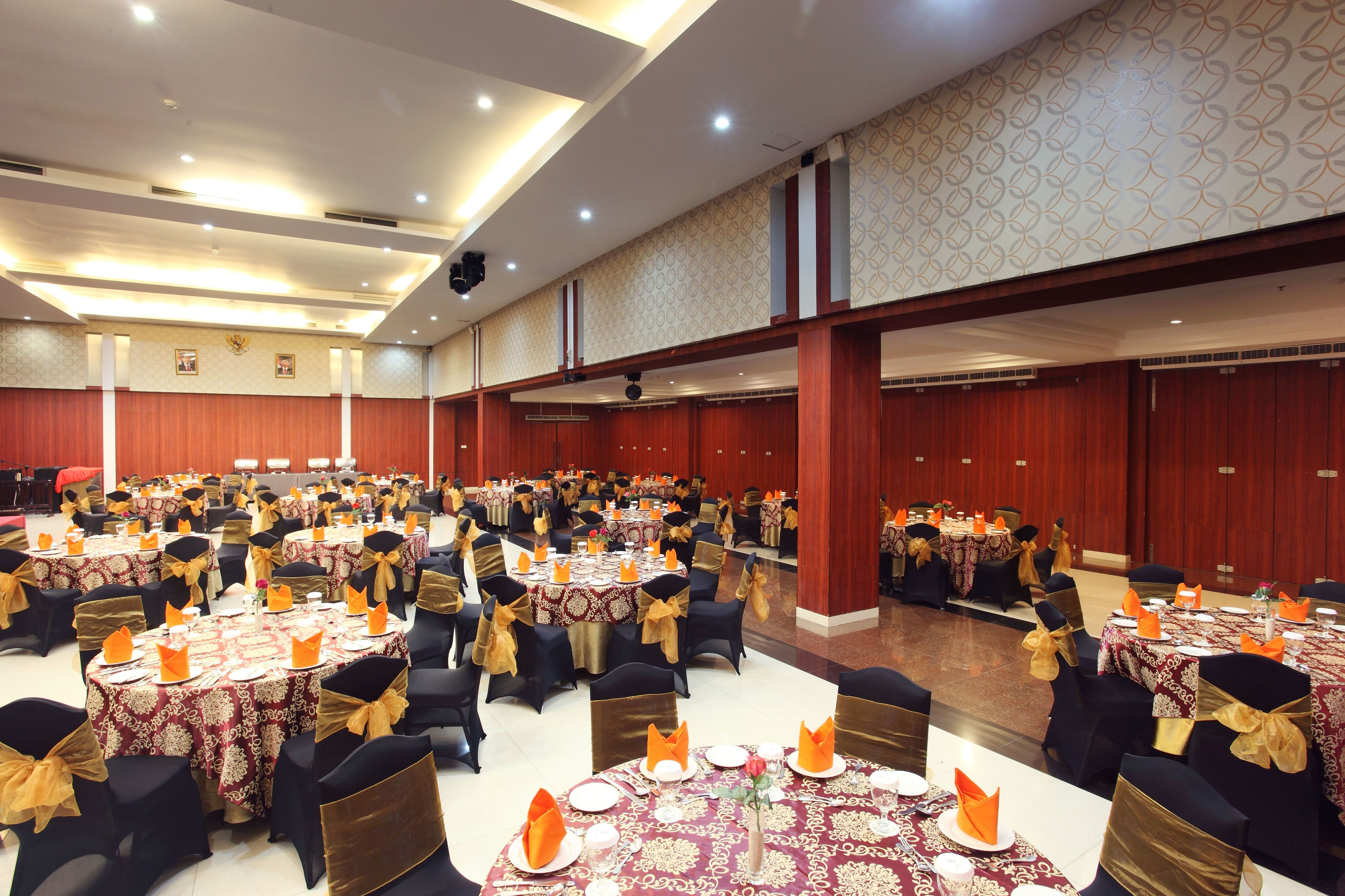Een feestzaal met meerdere ronde tafels, elk gedekt met witte tafellakens, gouden linten en oranje servetten.