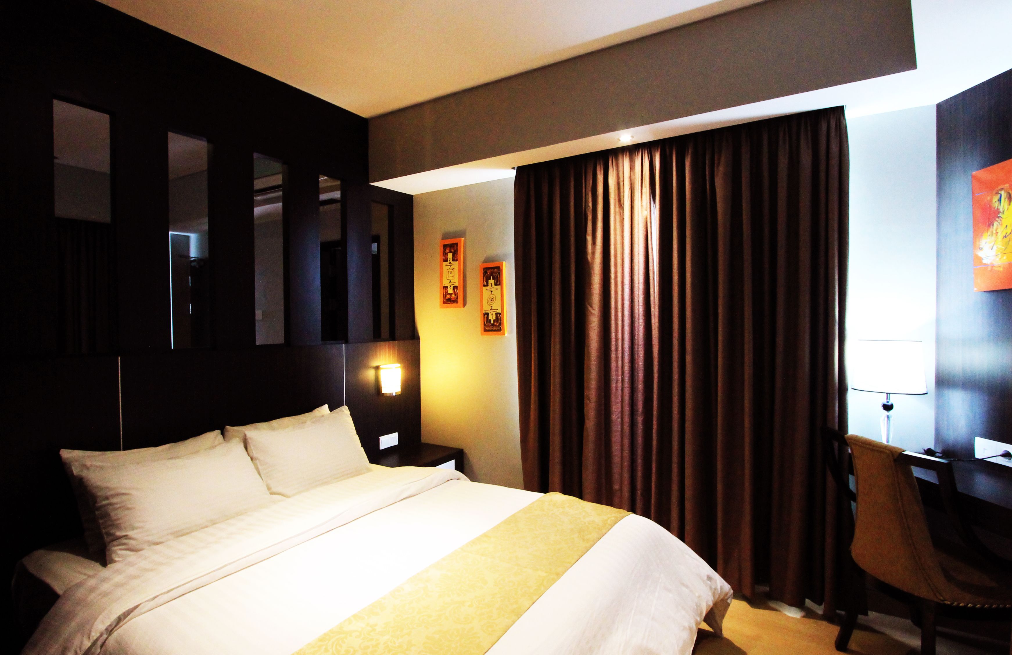 A hotel room with a large bed, a desk with a chair, a nightstand, and a wall-mounted lamp.