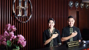 A reception desk with a 'RECEPTION' sign, a large metallic 'H' logo, and a vertical wooden panel background. - Grand Hatika Hotel (Tanjung Pandan)