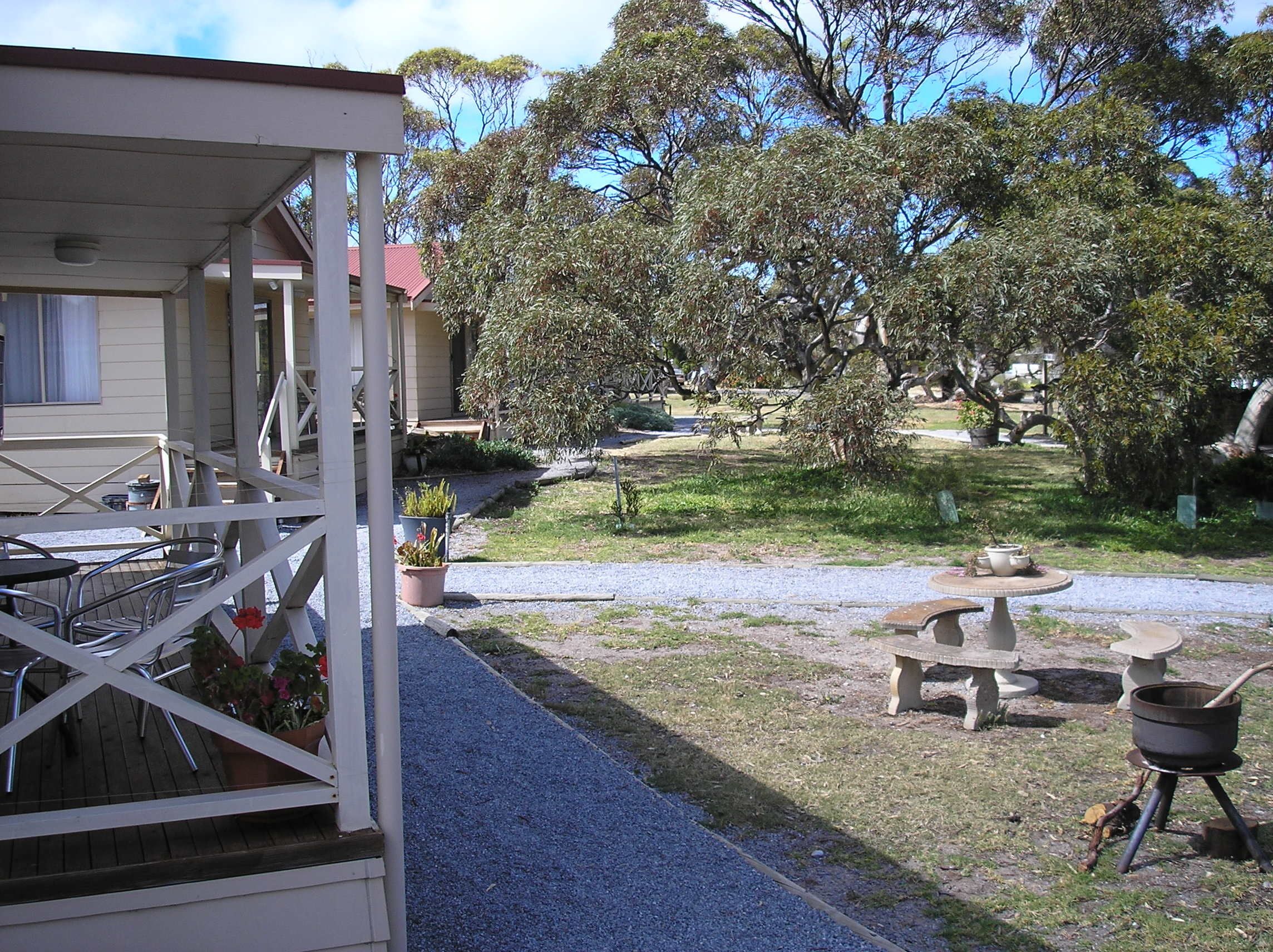 A covered outdoor area with a patio table and chairs, surrounded by potted plants and a gravel path leading to a building.