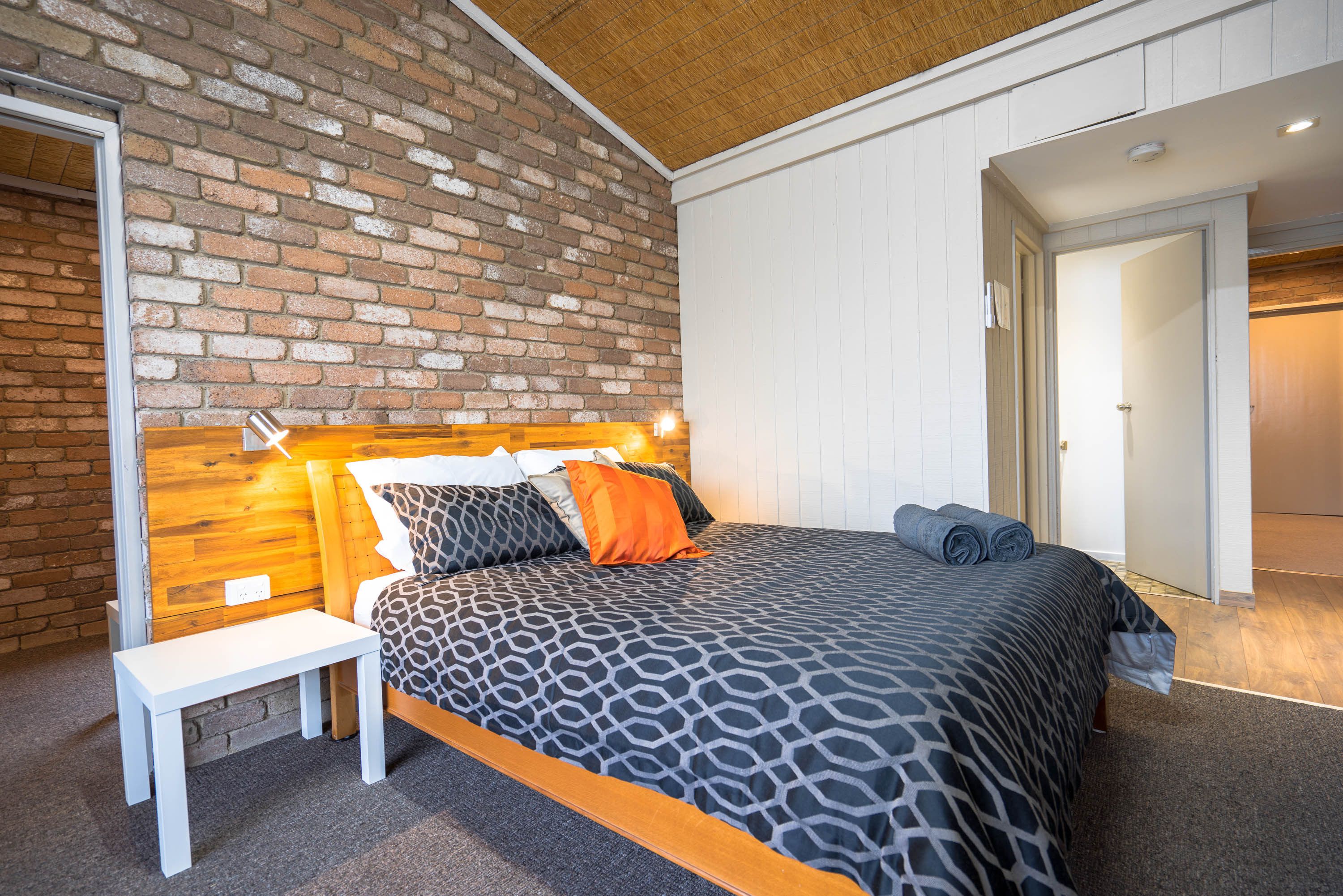 A bedroom with a brick wall, a wooden headboard, a patterned bedspread, and a white bench.