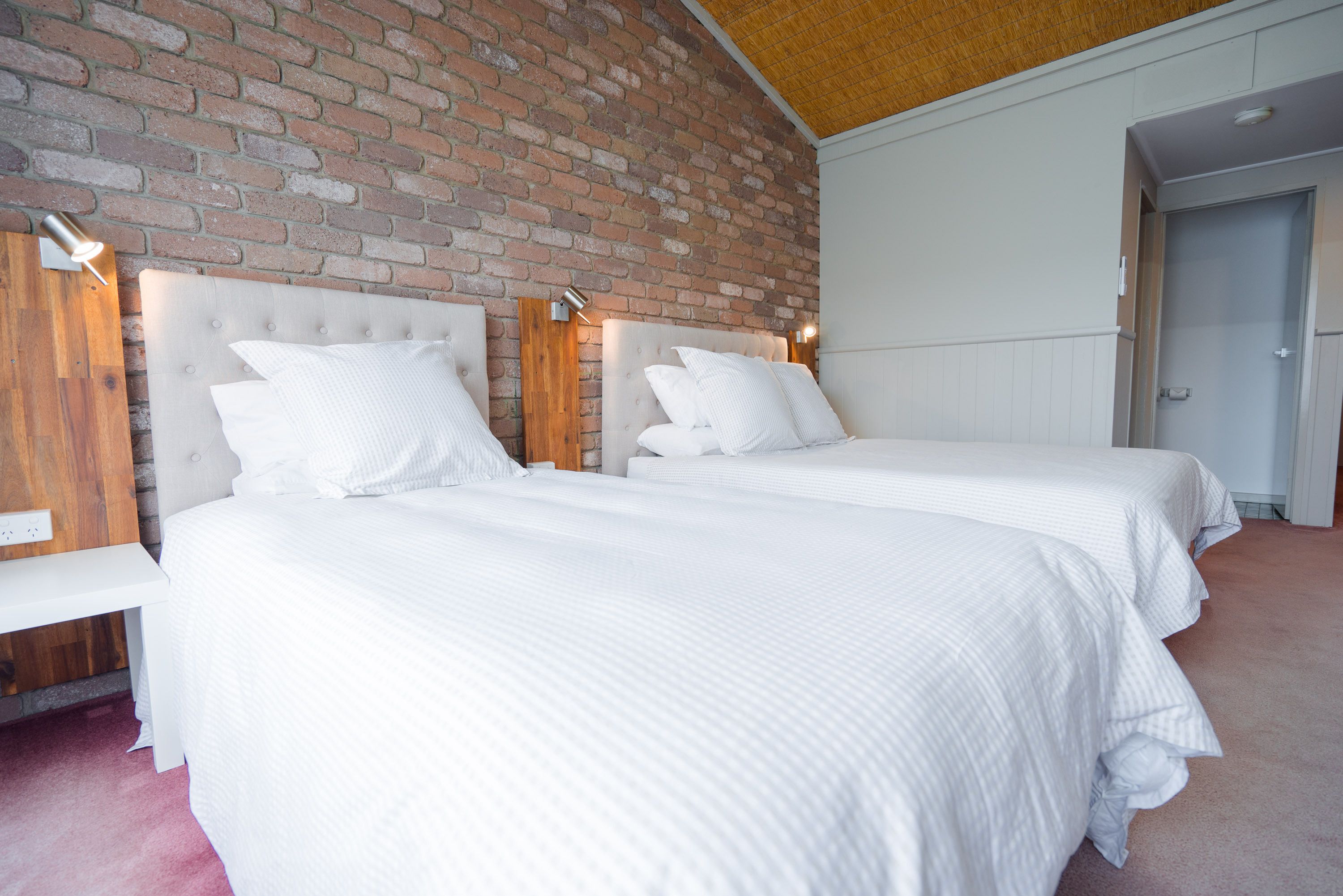 Hotellihuone, jossa on kaksi sänkyä, tiiliseinä ja puinen penkki.
