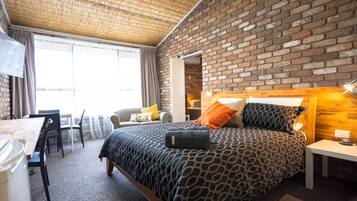 Une chambre d’hôtel avec un mur en briques, un lit avec une literie à motifs, un bureau avec une chaise et une grande fenêtre avec des rideaux.
