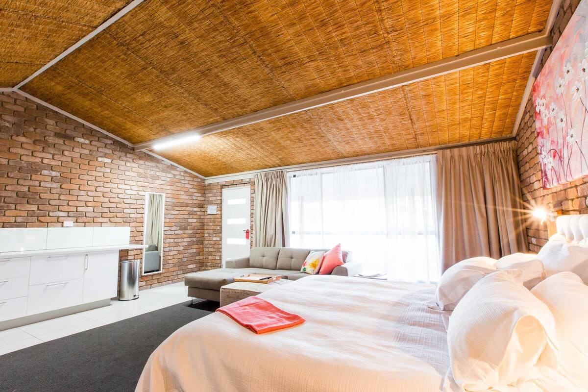 Una habitación de hotel moderna con pared de ladrillo, una cama grande con ropa blanca, un sofá y un área de comedor.