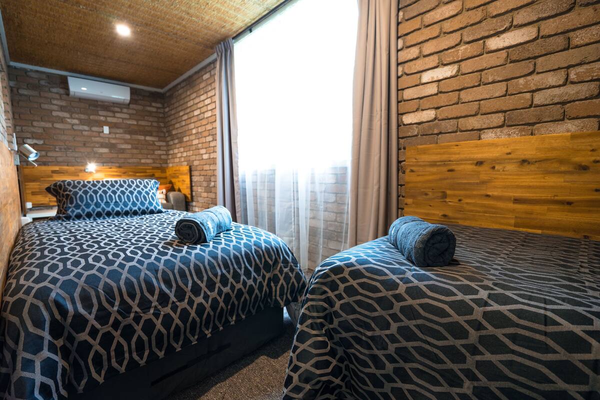Una habitación con dos camas, un muro de ladrillo, cabecero de madera y una ventana con cortinas.