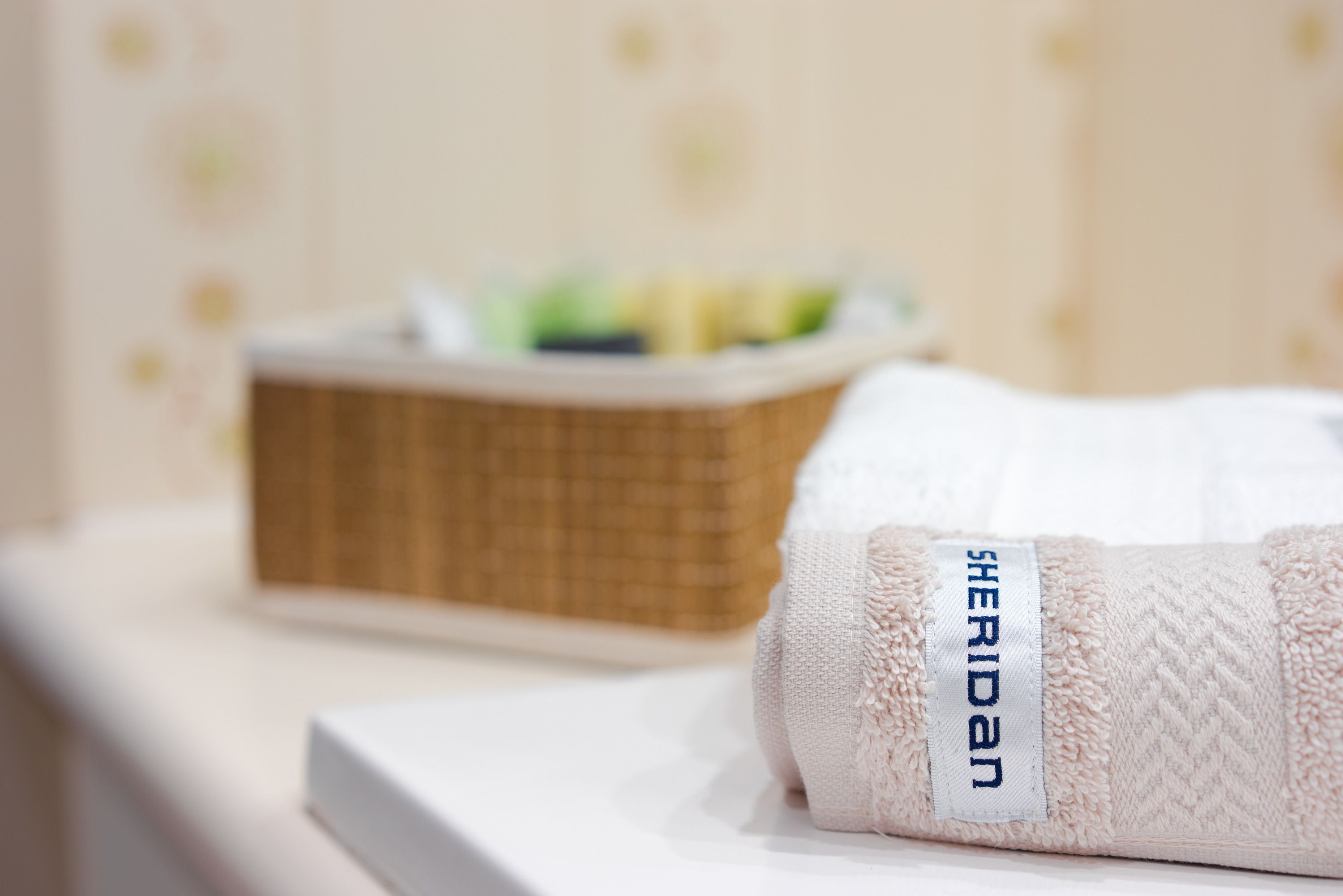 A neatly folded towel with a label reading "SHERIDAN" on a white surface, a wicker basket in the background, and a blurred wall with a floral pattern.