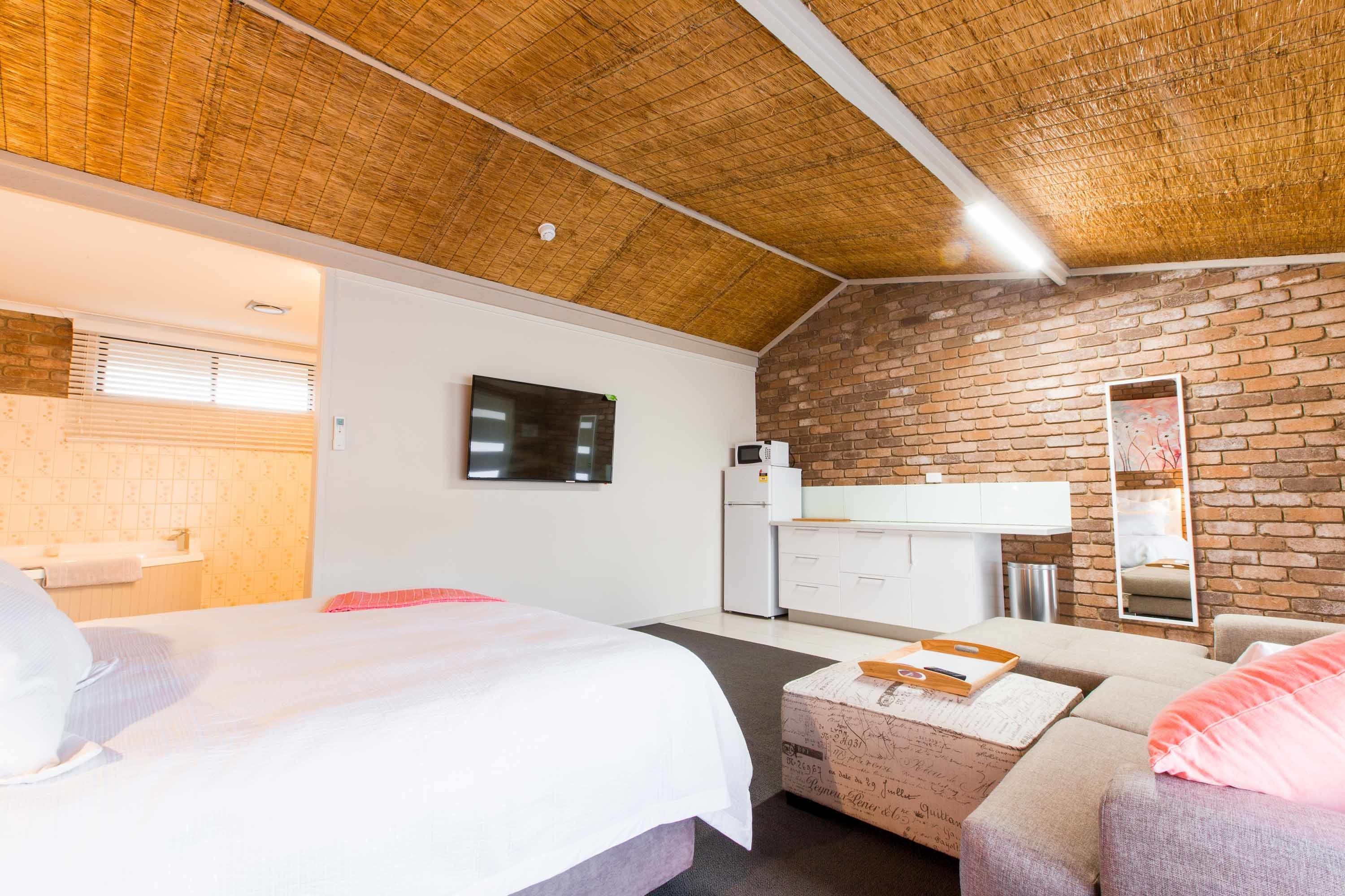 Une chambre avec un grand lit, un petit canapé, un plafond en bois, un mur en briques et un coin cuisine équipé d’un four à micro-ondes et d’un réfrigérateur.