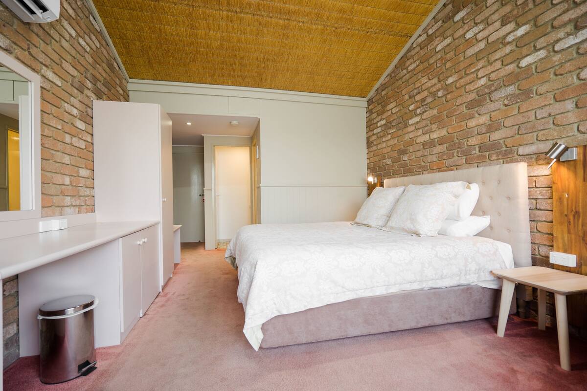 Un dormitorio con pared de ladrillo, una cama con ropa de cama blanca, una mesita de noche y un cubo de basura pequeño.