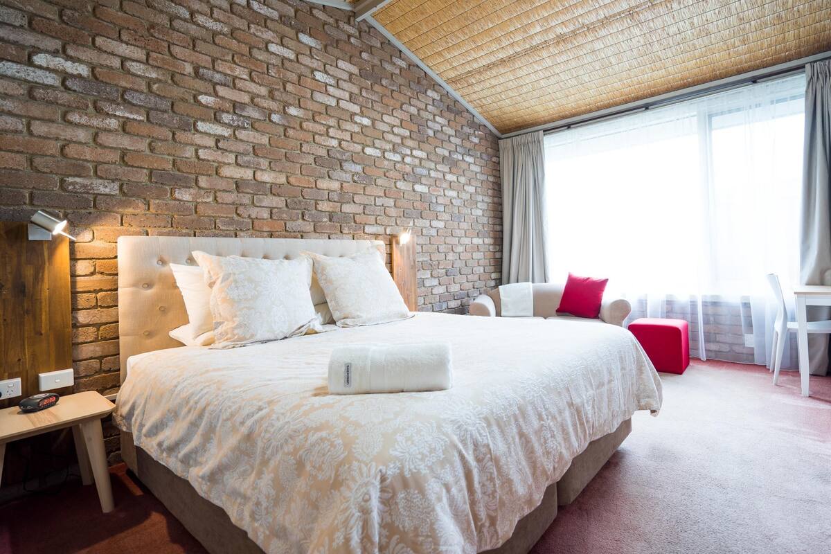 Habitación de hotel con pared de ladrillo, cama con ropa blanca, mesita de noche de madera y ventana con cortinas.