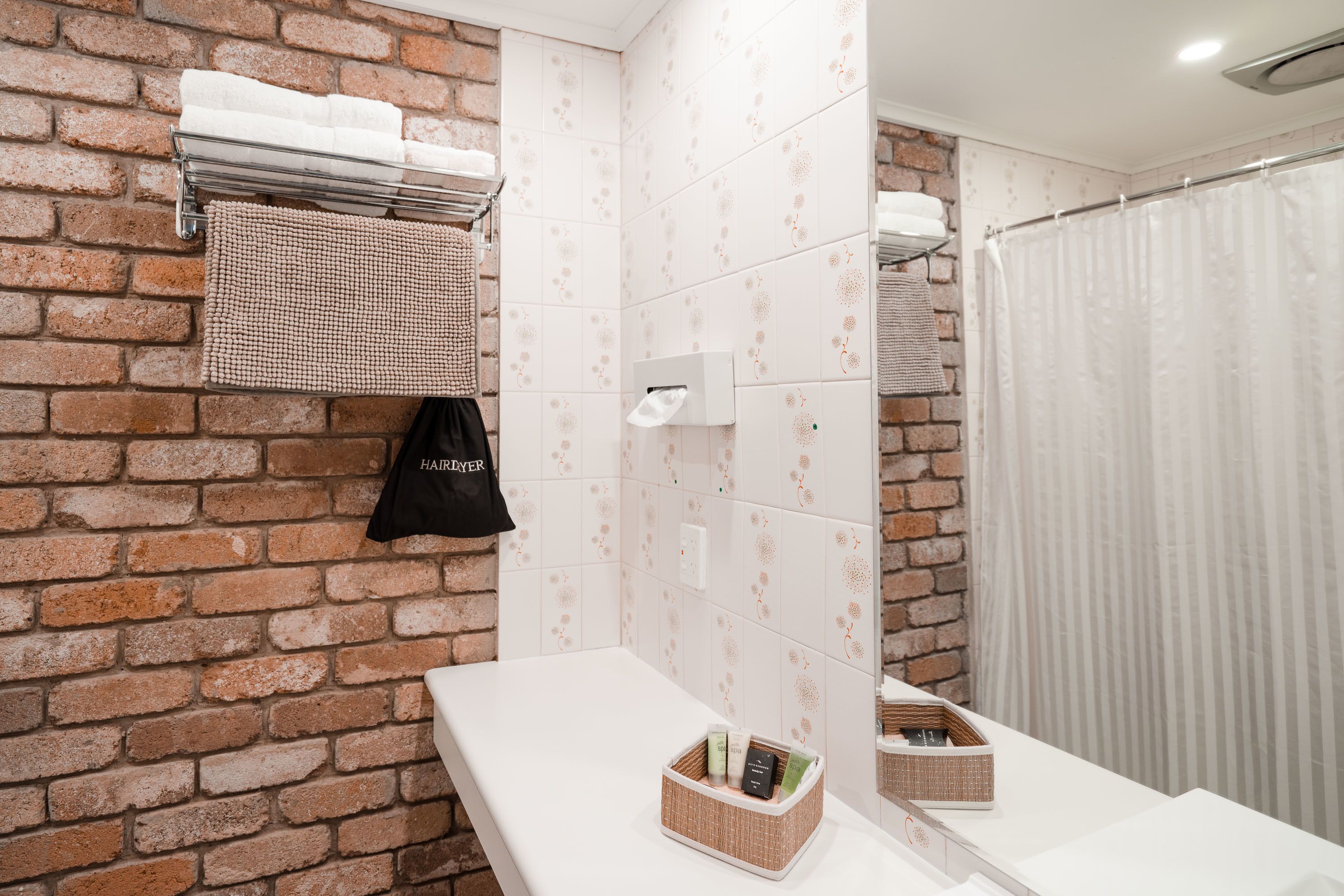 Un baño con pared de ladrillo, encimera blanca, toallero, cortina de ducha y espejo.