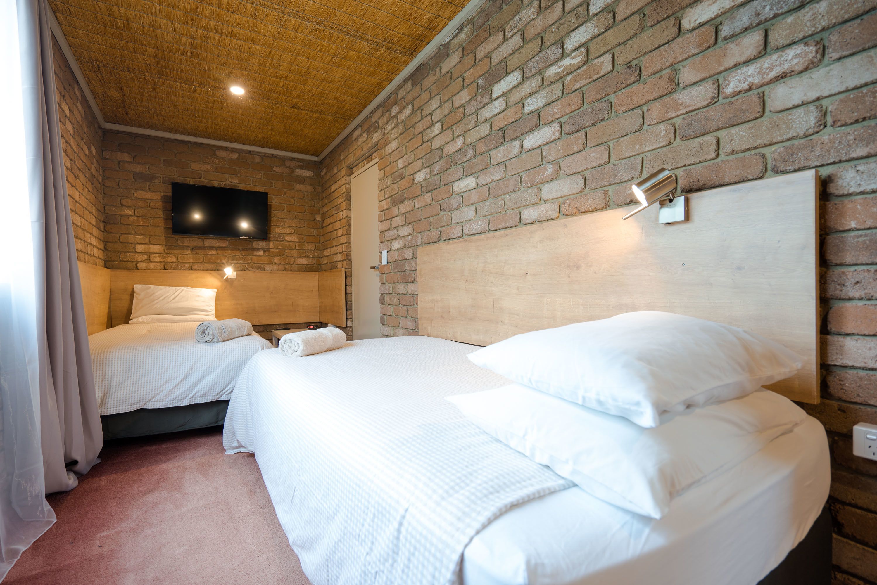 Habitación de hotel con dos camas, una pared de ladrillo, un televisor de pantalla plana montado sobre las camas y un cabecero de madera.