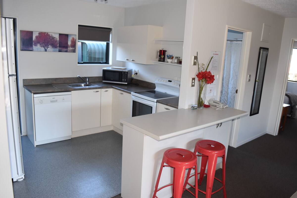 Una cocina moderna con armarios blancos, microondas, fregadero y una pequeña isla con dos taburetes rojos.