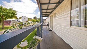 A covered porch with a wooden deck, a window with curtains, and a view of other RVs in the background. - Warragul Gardens Holiday Park (Warragul)