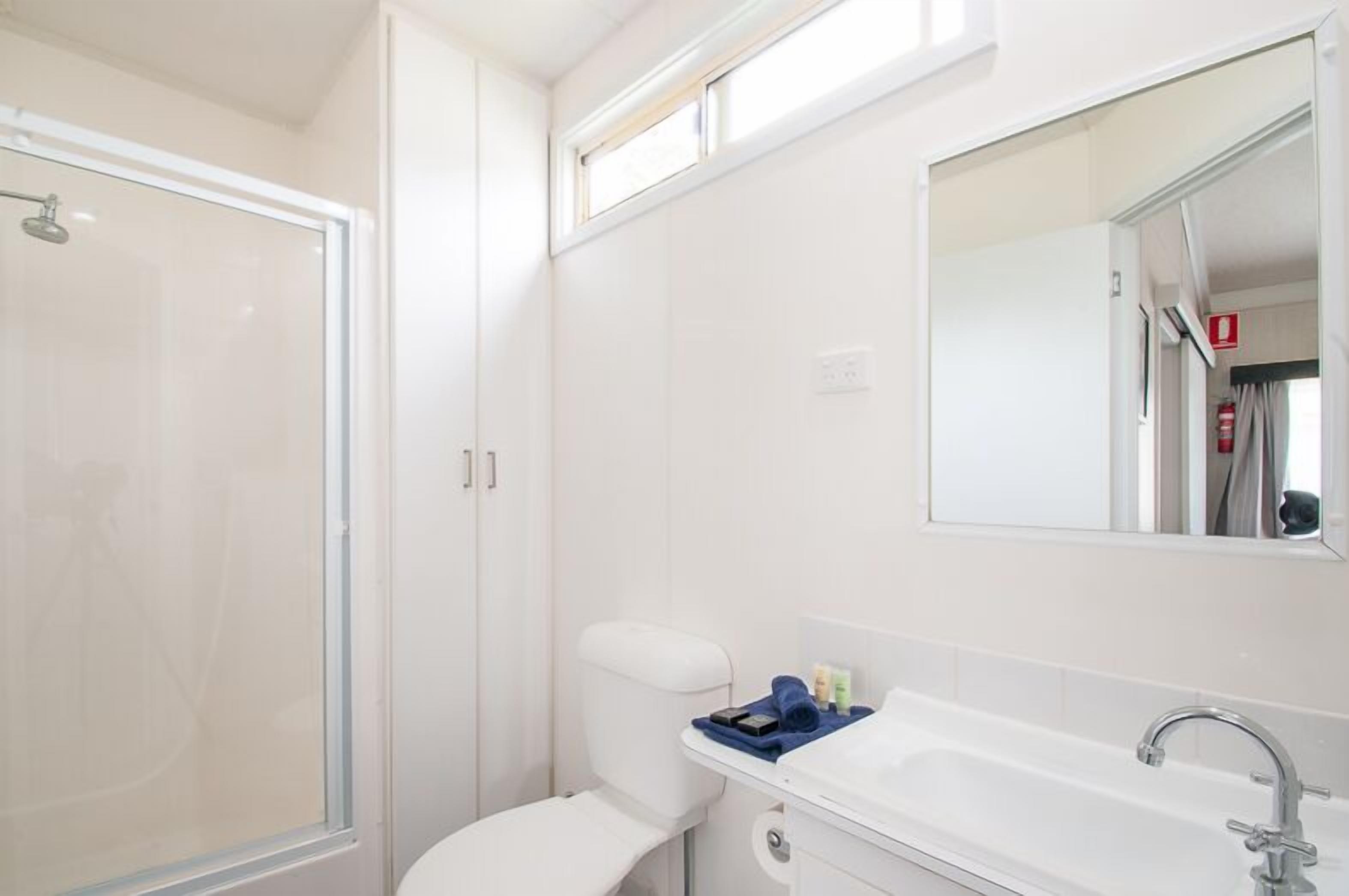 Un baño con cabina de ducha de vidrio, inodoro blanco, lavamanos con espejo y un pequeño estante con artículos de aseo.
