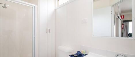 Un baño con cabina de ducha de vidrio, inodoro blanco, lavamanos con espejo y un pequeño estante con artículos de aseo.
