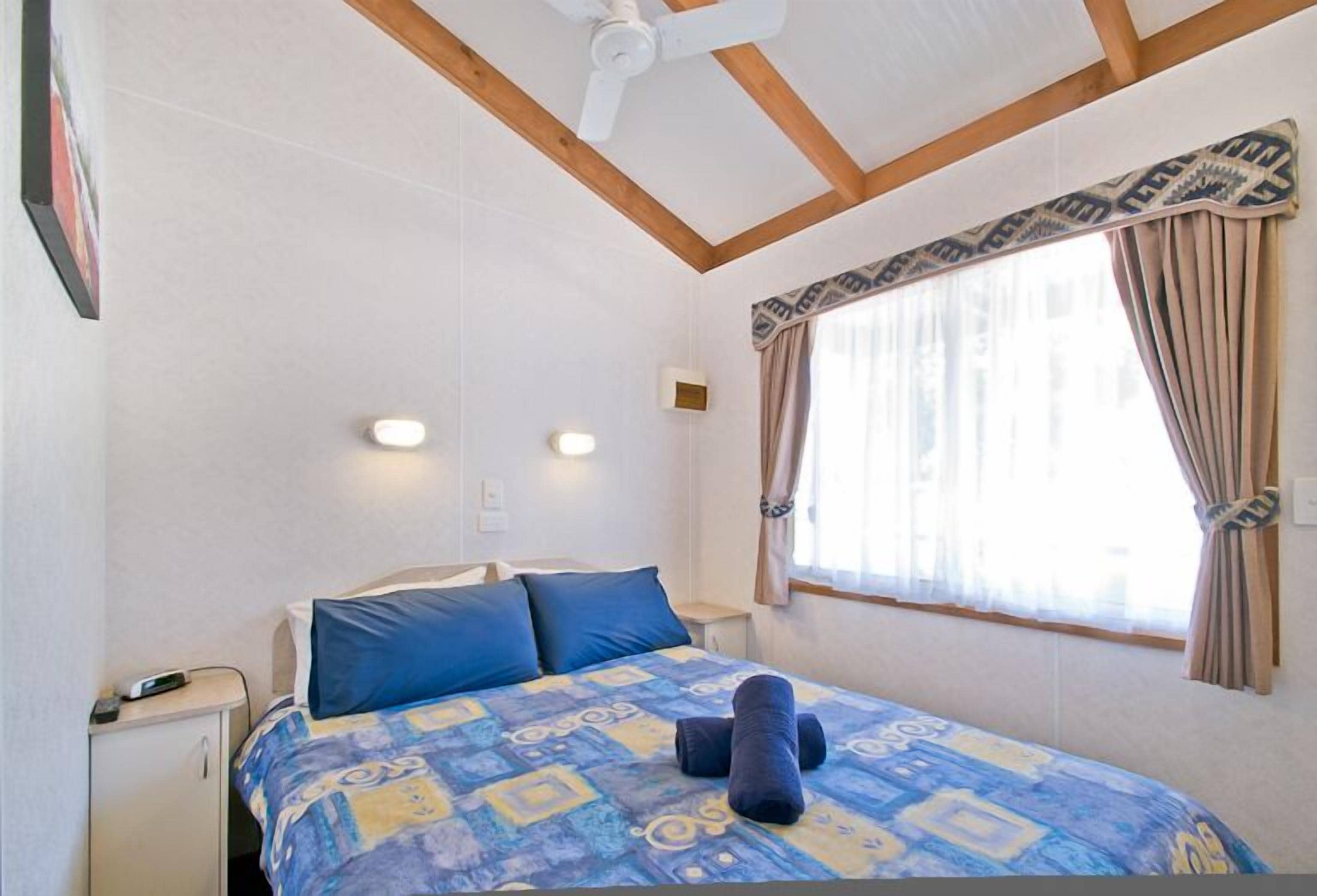A bedroom with a bed, a nightstand, a ceiling fan, a window with curtains, and a wall-mounted light.