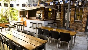 An outdoor dining area with a long wooden table and multiple chairs, a bar area with stools, and a brick building in the background. - Queens Arms Hotel (Roma)