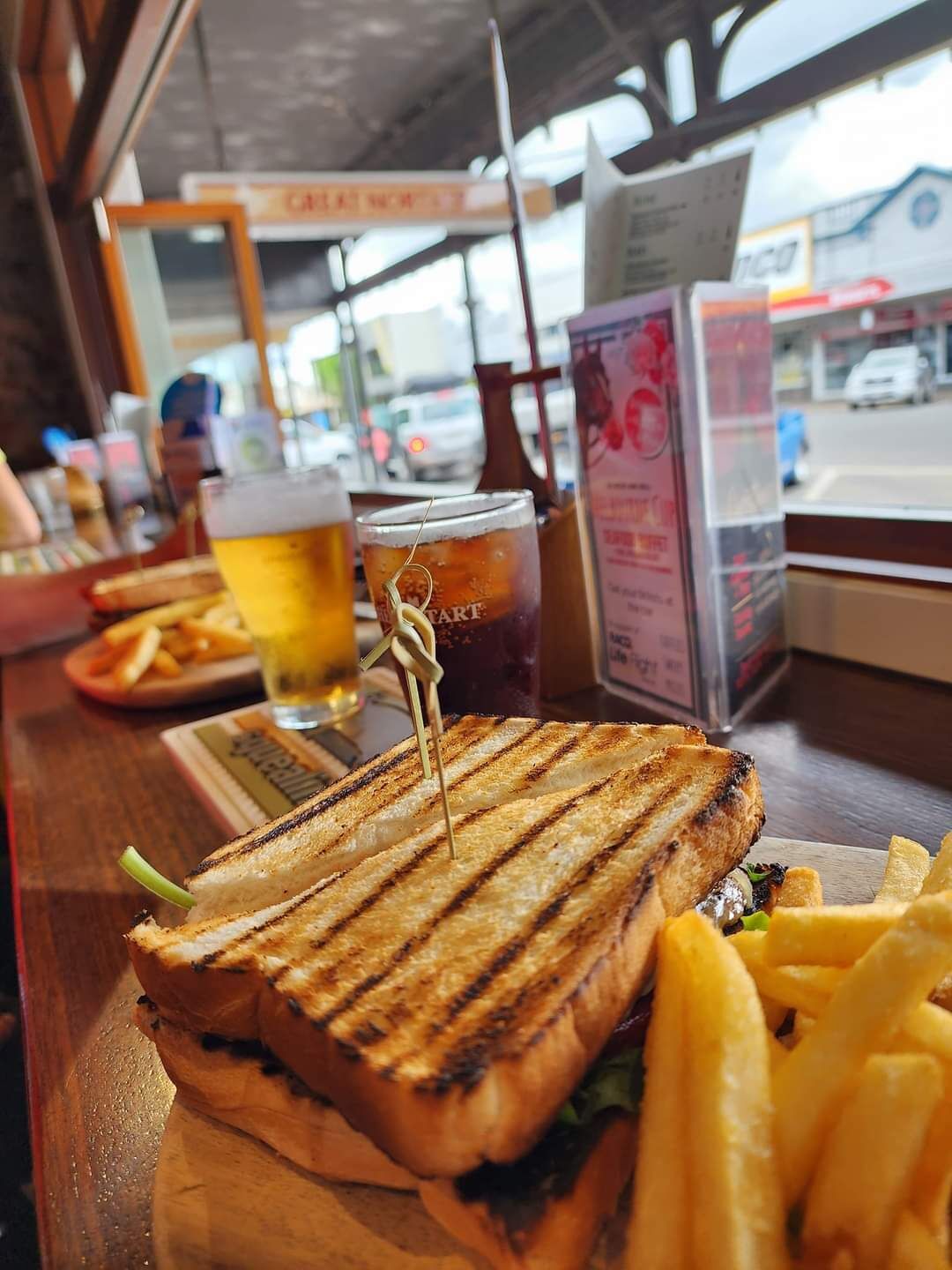 En smörgås med en tandpetare i, ett glas öl och pommes frites på ett bord.