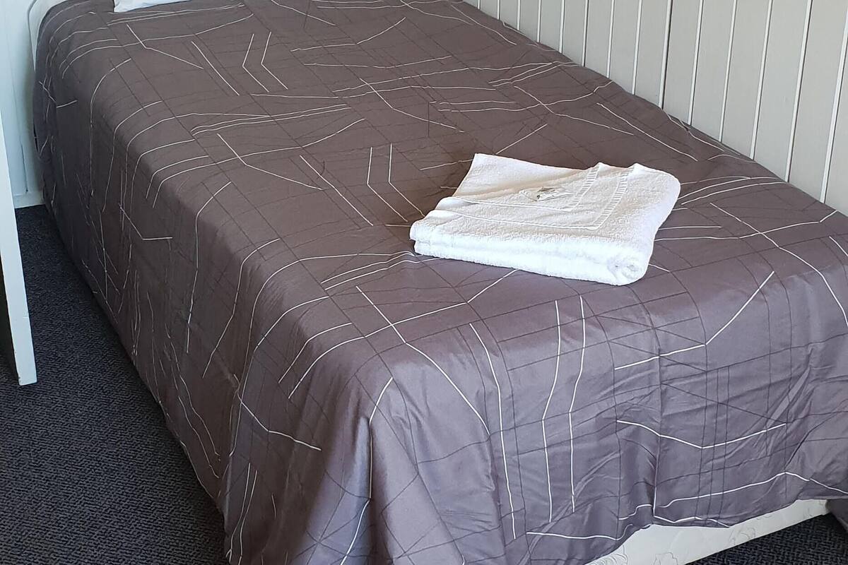 A single bed with a brown bedspread and a white pillow, a folded white towel, and a dark carpeted floor.