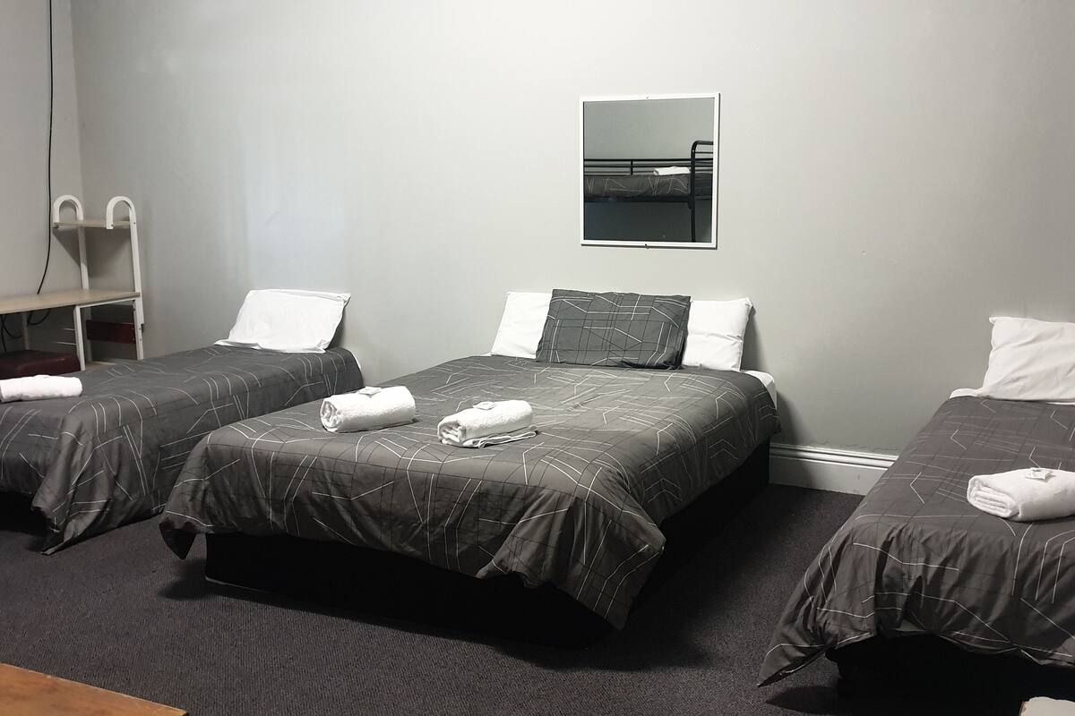 A room with three beds, each with a dark patterned bedspread and white pillows. There is a small table with a chair, a mirror on the wall, and a carpeted floor.