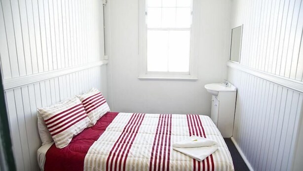 A small, white-walled bedroom with a single bed, a bedside table, and a window.