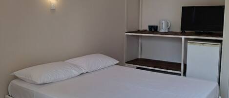 Habitación individual con una cama blanca, un escritorio pequeño con televisor y una lámpara de pared.