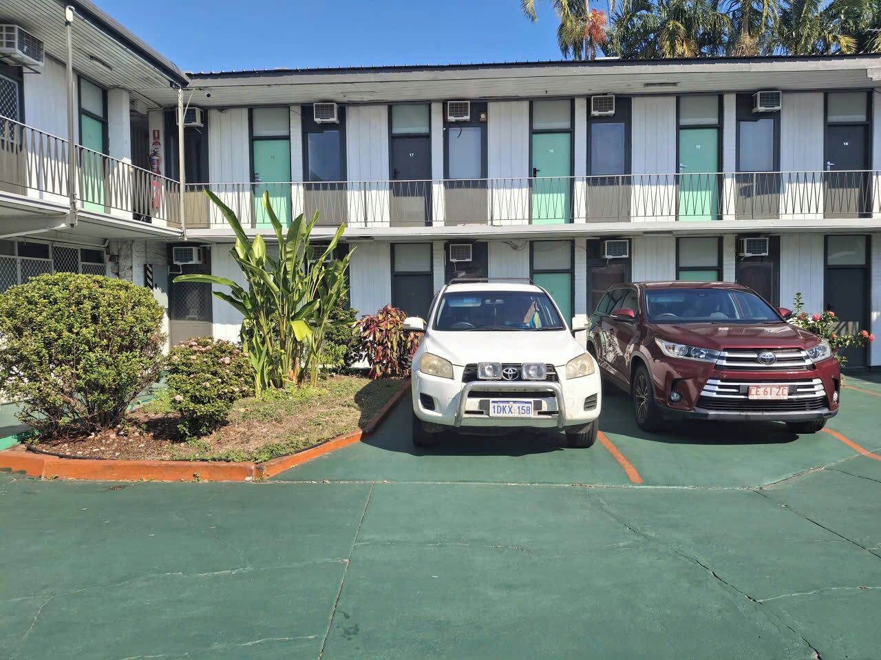 A multi-story building with a parking area in front. There are two cars parked, one white and one red. The building has multiple windows and balconies.