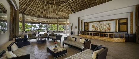 A spacious lounge area with multiple sofas and armchairs, a large artwork on the wall, and a thatched roof ceiling.