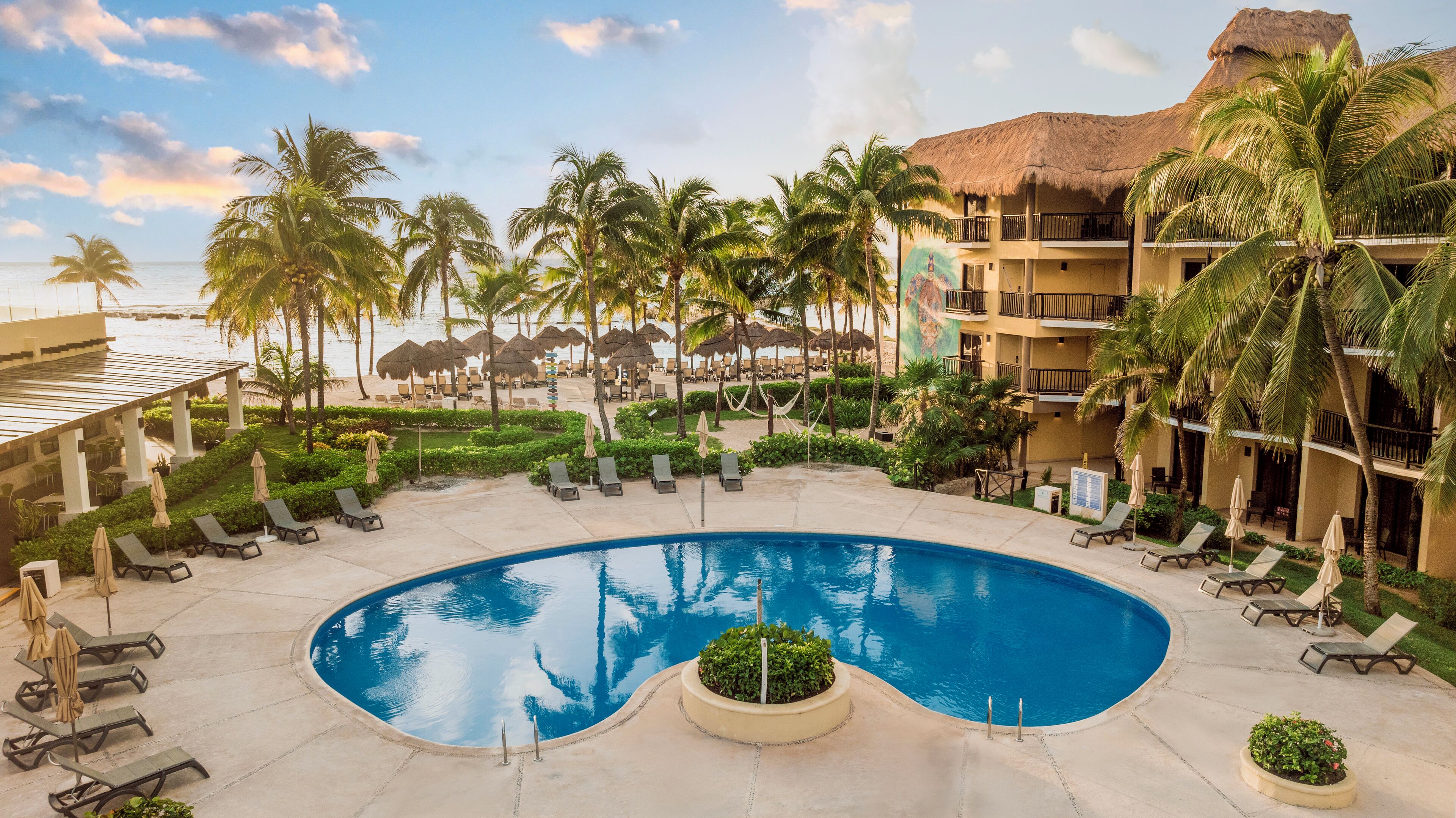 Zona de piscina en un resort con sillones de descanso, palmeras y un edificio con techo de paja.