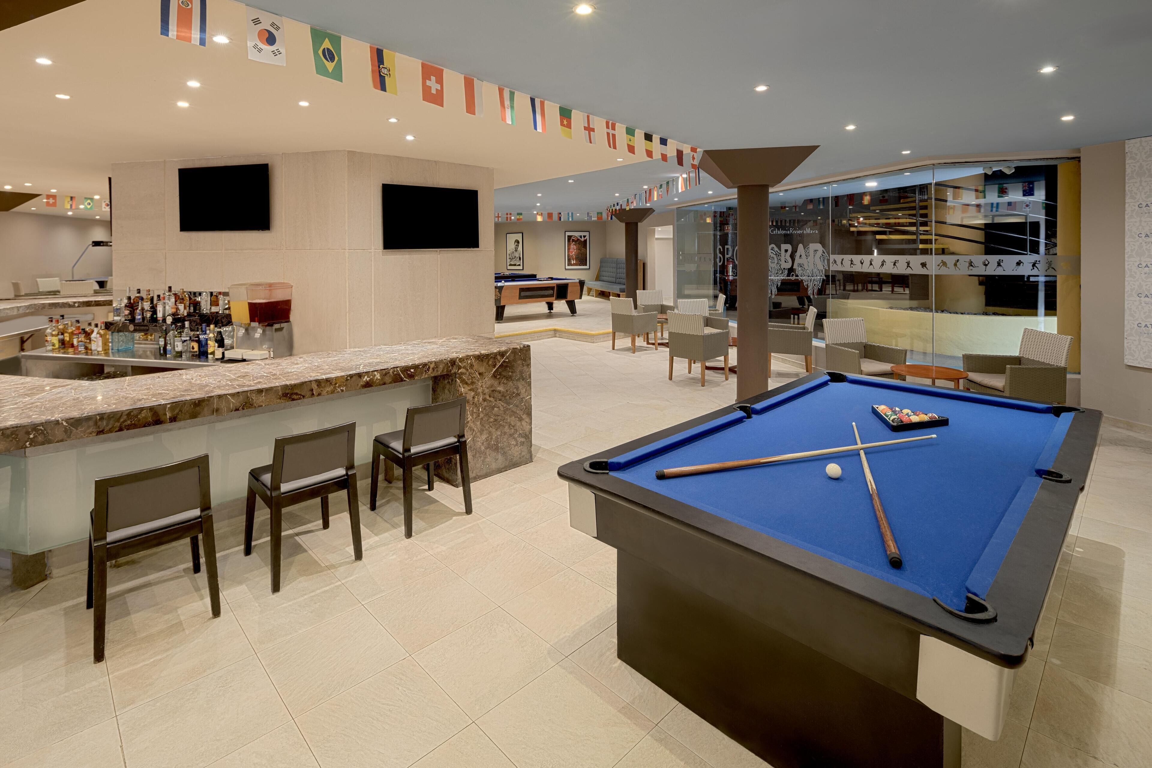 A pool table with pool cues and a billiard ball, surrounded by bar stools and a bar area with bottles.