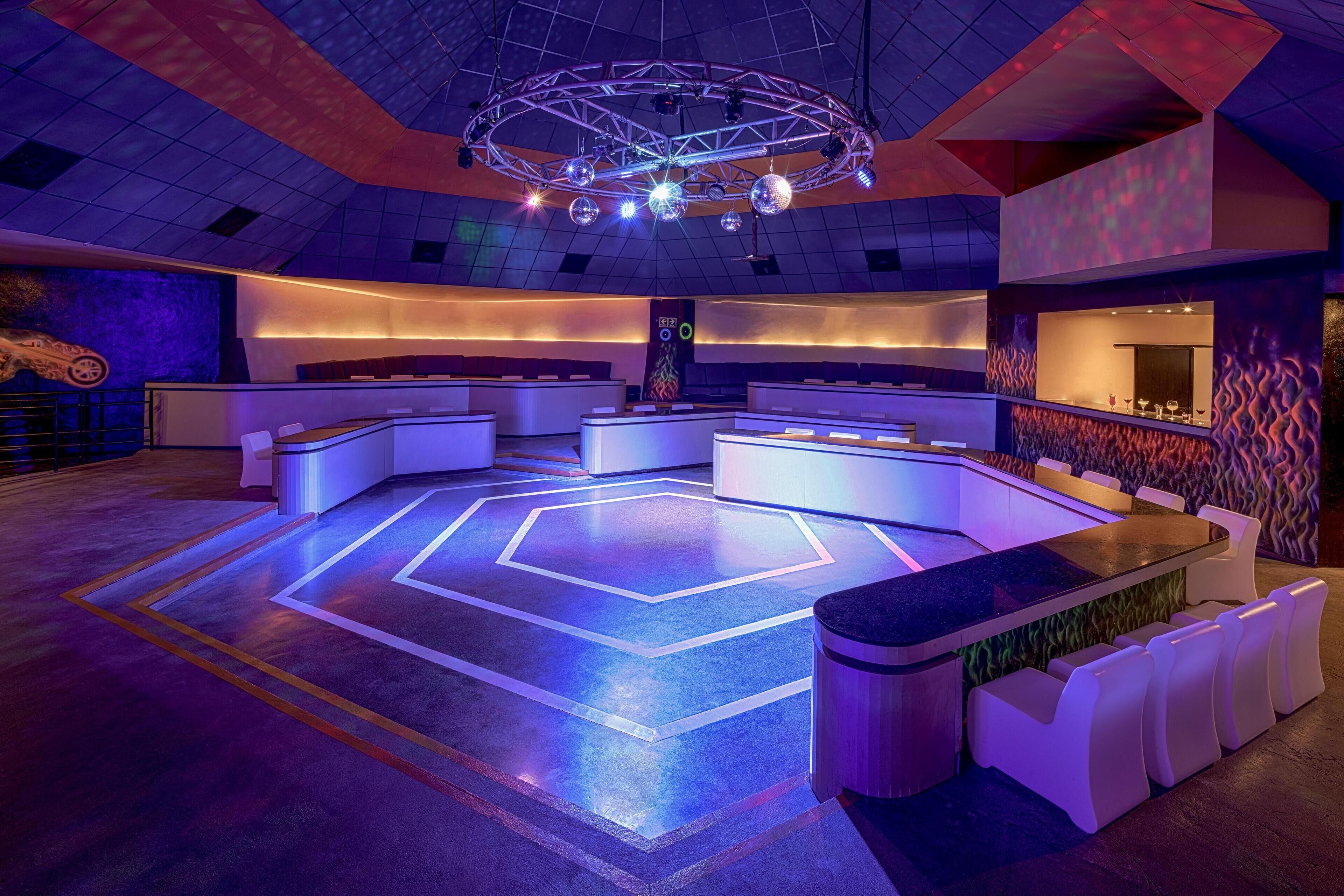A modern lounge area with a central seating arrangement, a bar area with a unique wavy wall design, and a ceiling with a geometric light fixture.