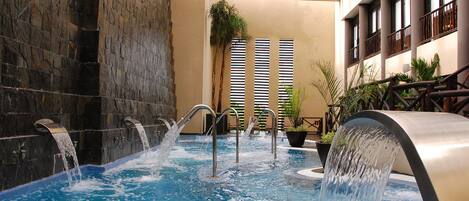 An indoor swimming pool with multiple water jets, surrounded by stone walls and potted plants.