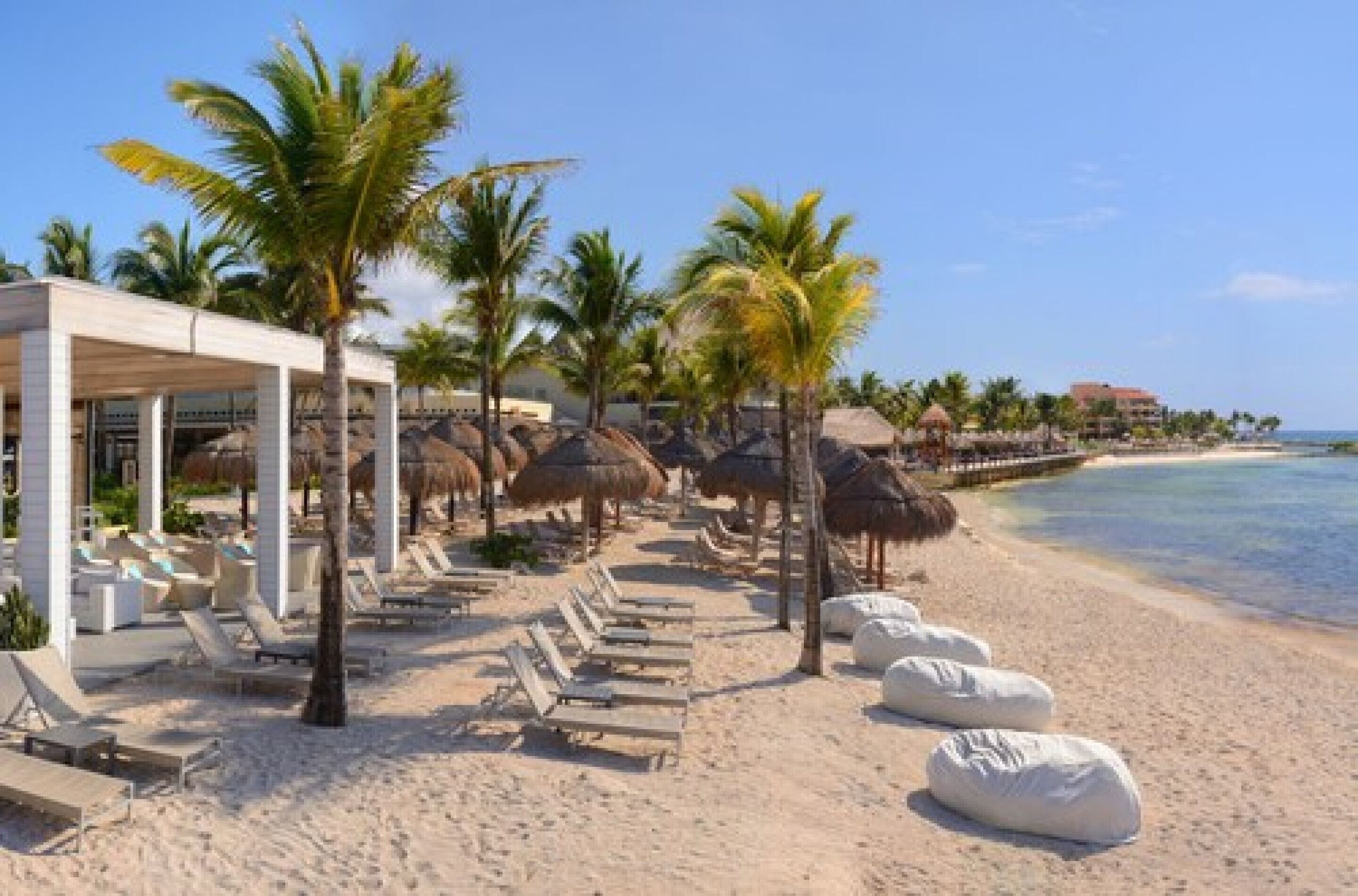 Un resort en la playa con sillas de descanso, palmeras y un edificio con zona cubierta.