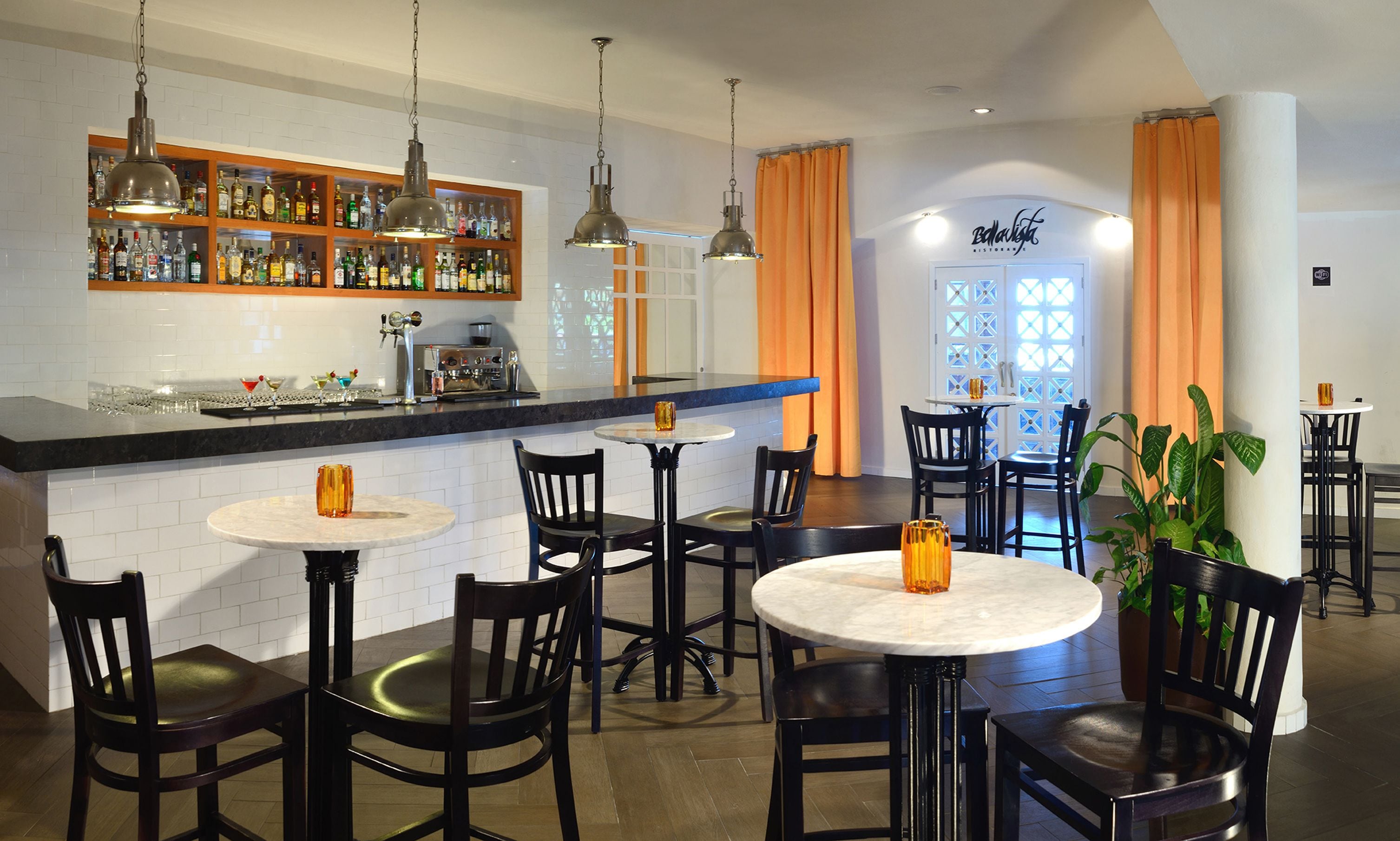 A bar area with a counter, shelves of bottles, and hanging lights. There are tables and chairs arranged for seating.