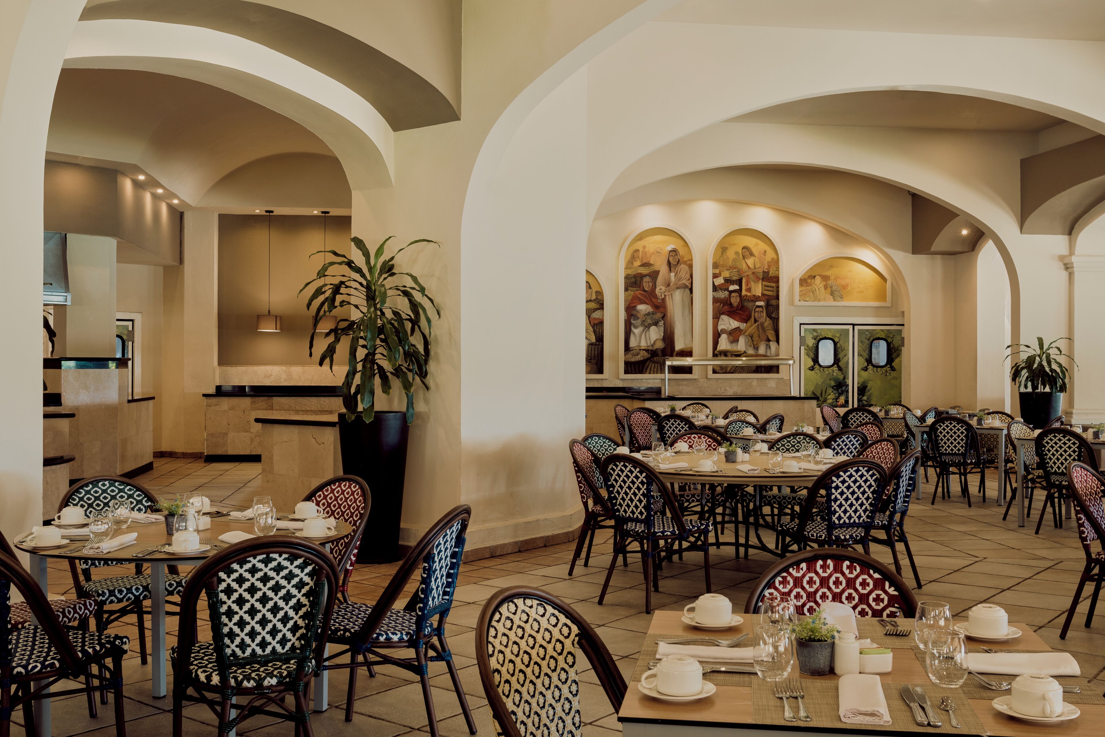 A dining area with tables and chairs, a large potted plant, and a mural on the wall.