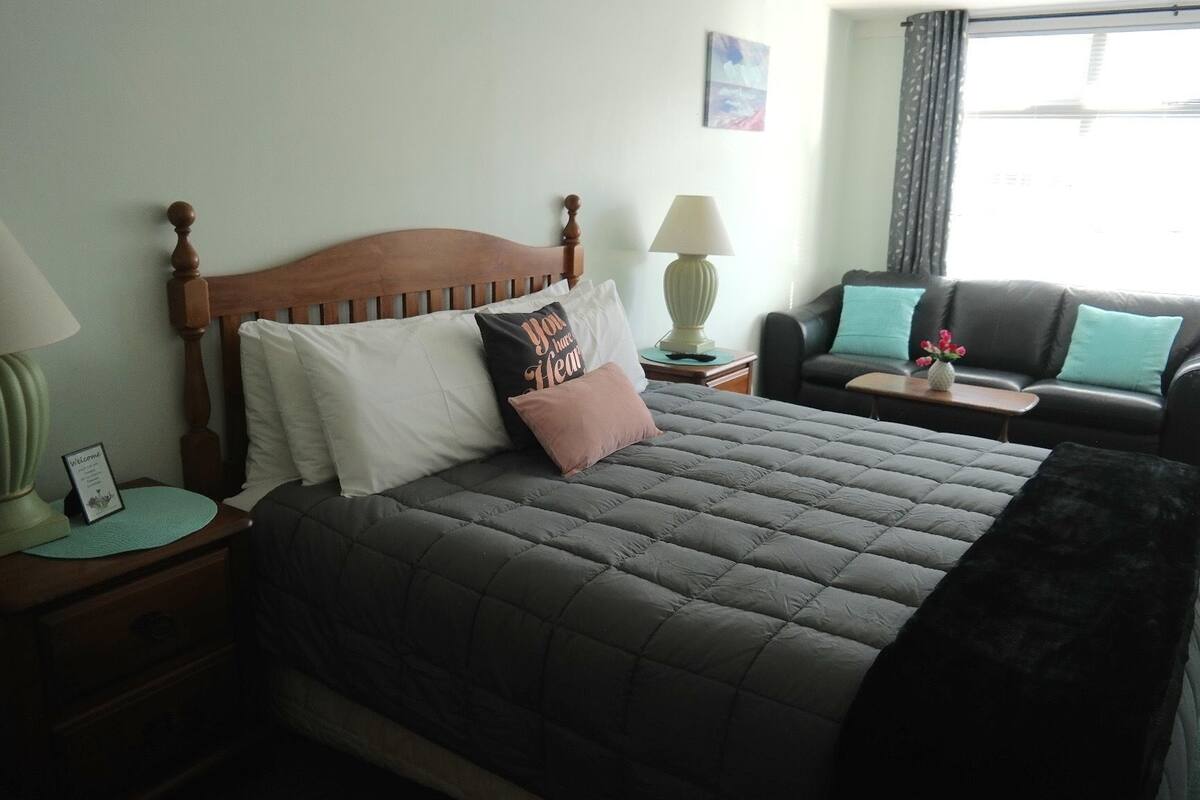 A neatly made bed with a dark quilt, a wooden headboard, and several pillows, including one with text. There is a bedside table with a lamp and a small sign, and a chair with a cushion.