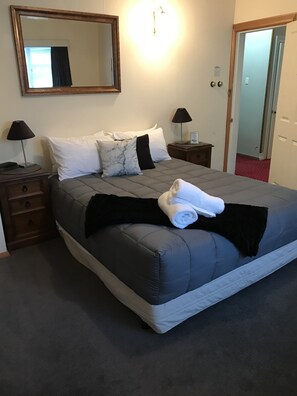 Un lit bien fait, avec un matelas gris et du linge de lit blanc, deux tables de chevet avec des lampes, un grand miroir au-dessus du lit et une porte sur la droite.